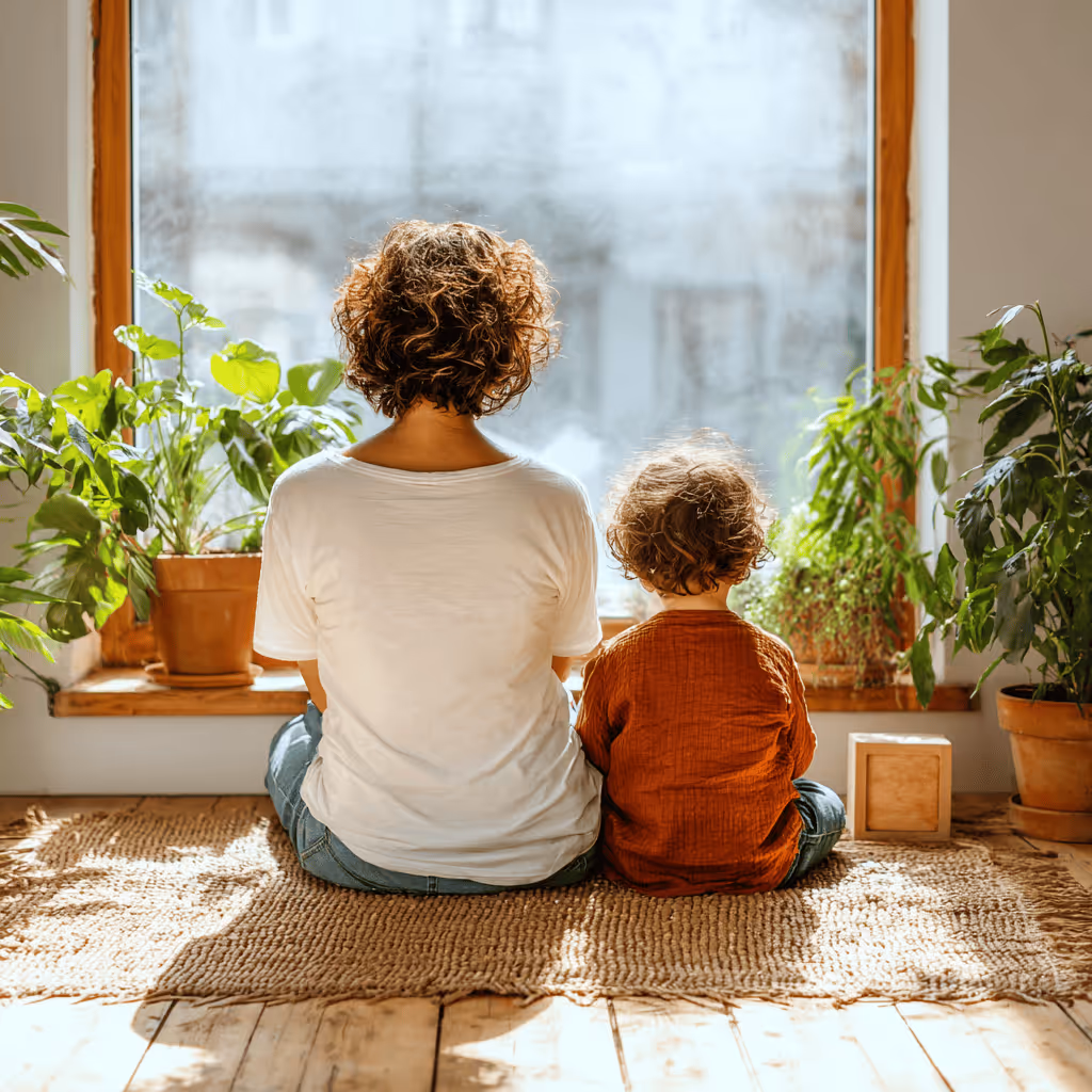 Adult and child with curly hair sitting on a woven rug facing a sunlit window surrounded by potted plants.