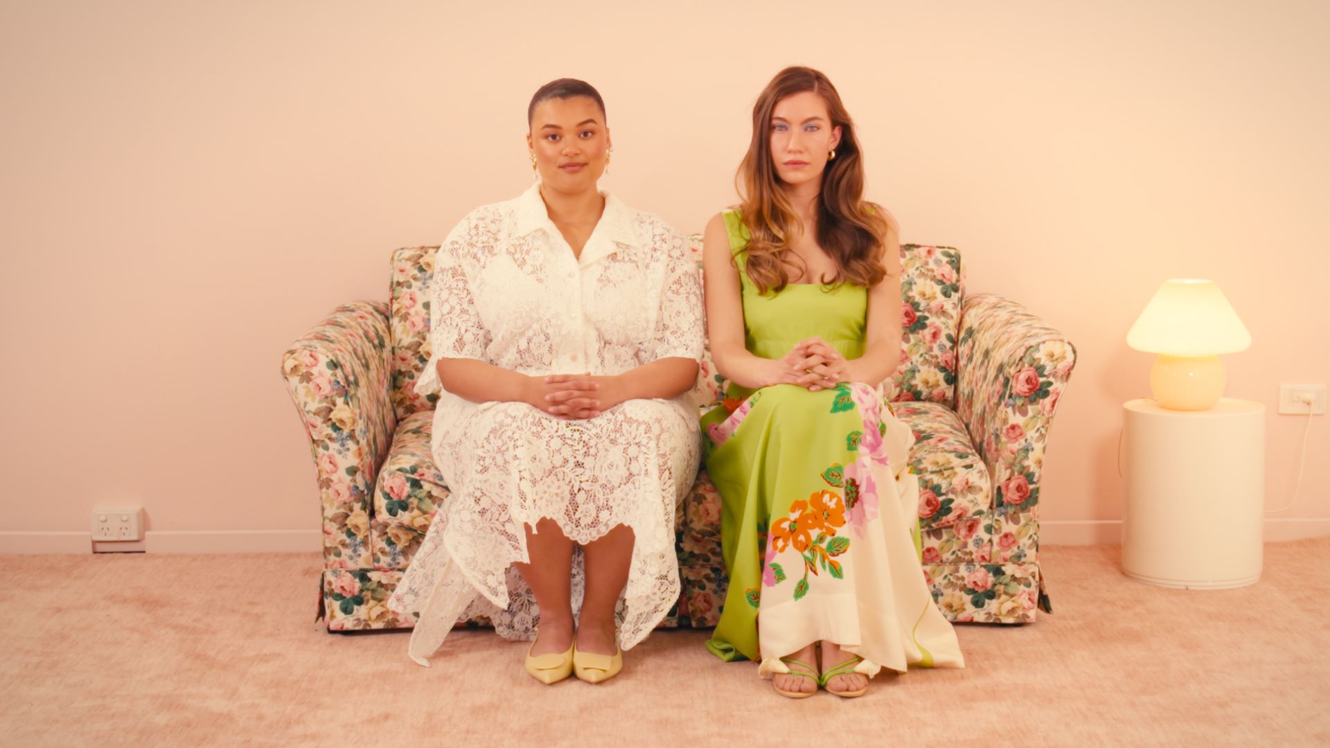 Two models sit on a floral couch in a pink pastel living room. 