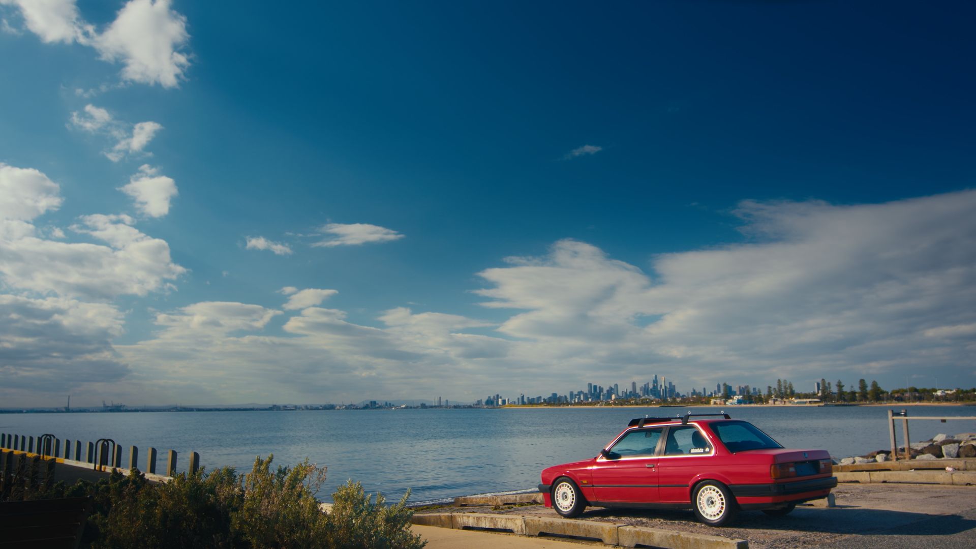 A red car parked by the ocean with a city skyline in the background