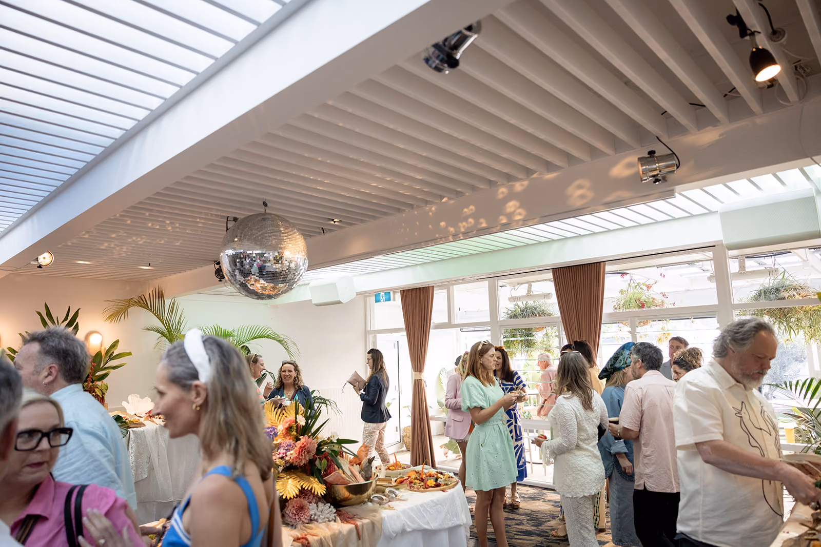 People socializing by flower-decorated buffet tables under a disco ball in a sunny event space.