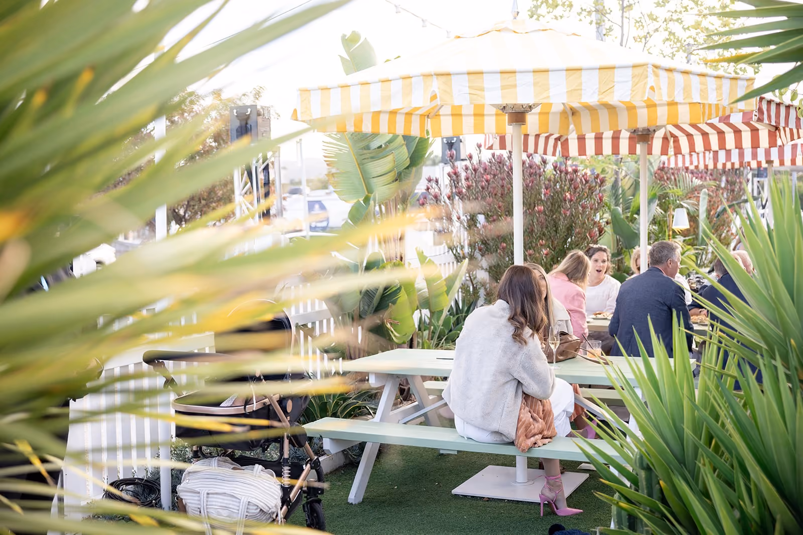 People sitting and chatting under striped umbrellas at picnic tables in a green garden setting.