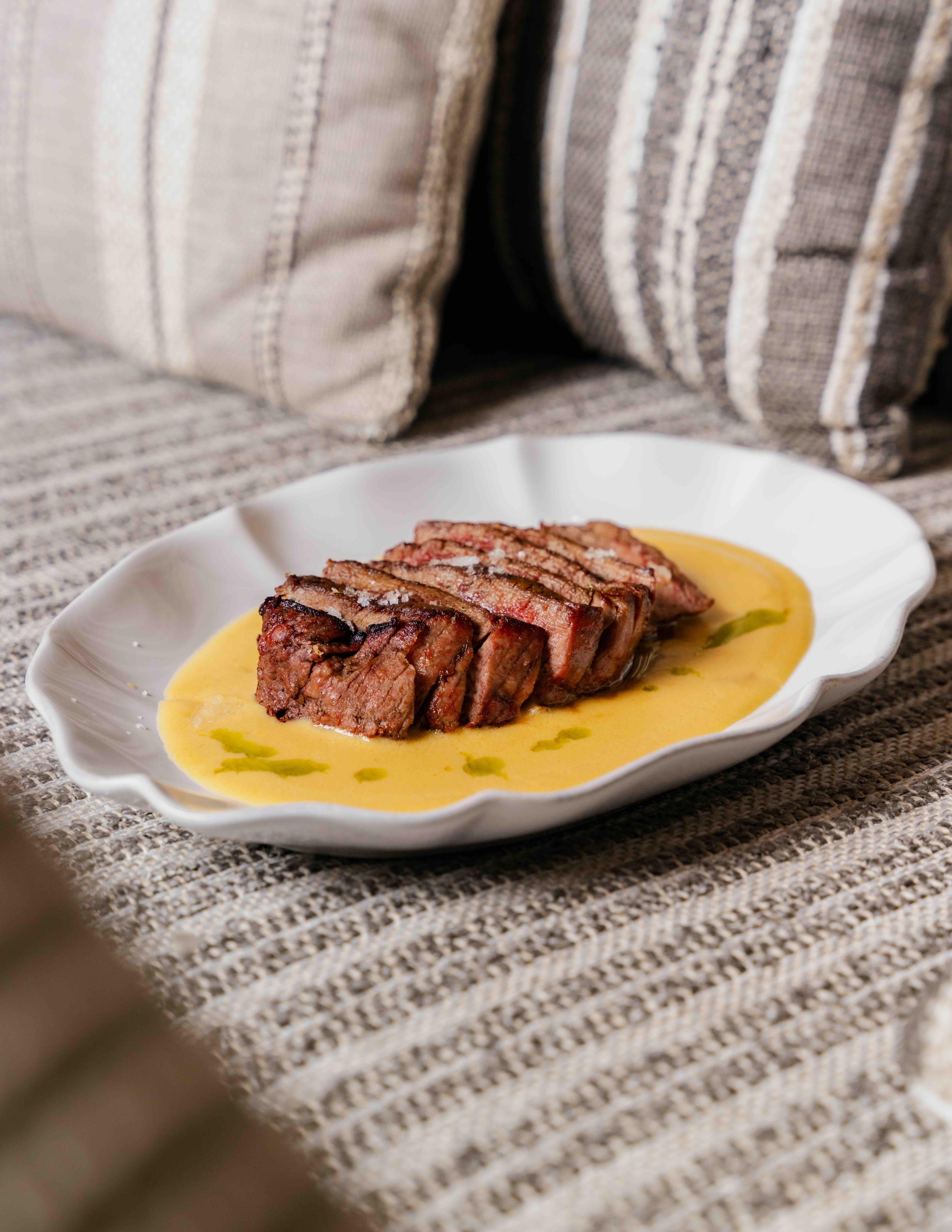 Sliced grilled steak on a white plate with creamy yellow sauce and green oil drizzles.