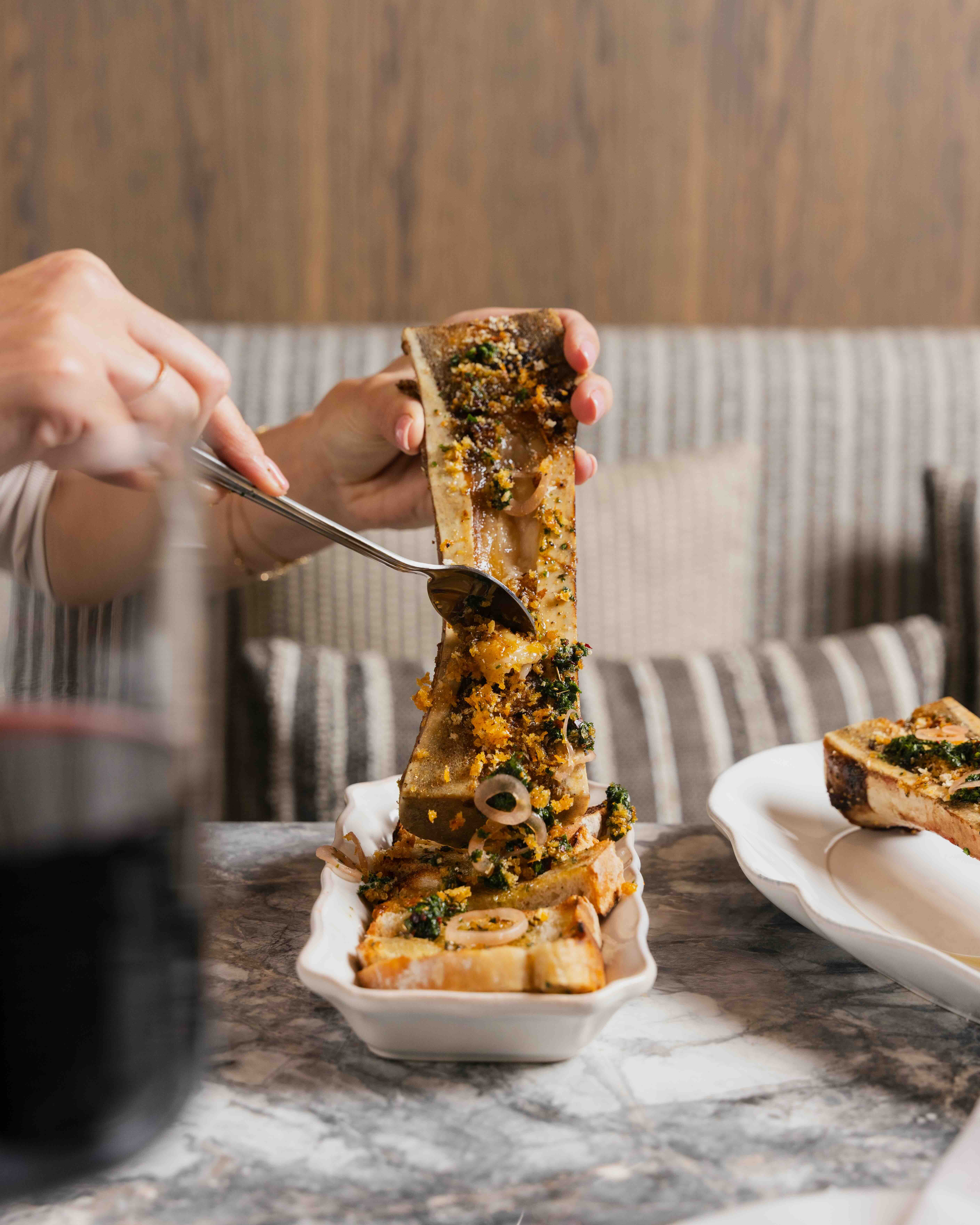 Hands serving roasted bone marrow with herbs and onions on toasted bread in a white dish.