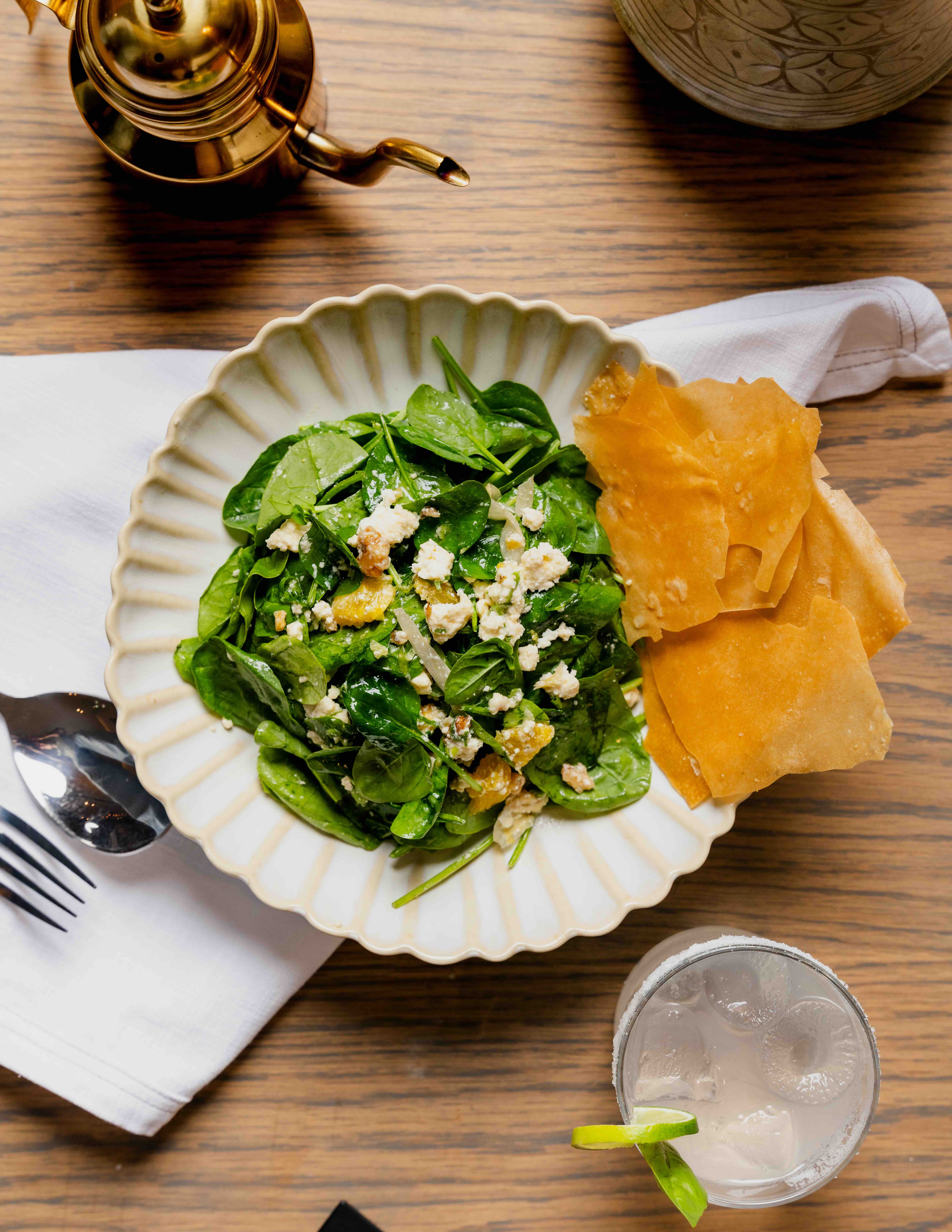 Plate of spinach salad with feta and orange pieces, crispy chips, drink with lime, and brass teapot on wood table.