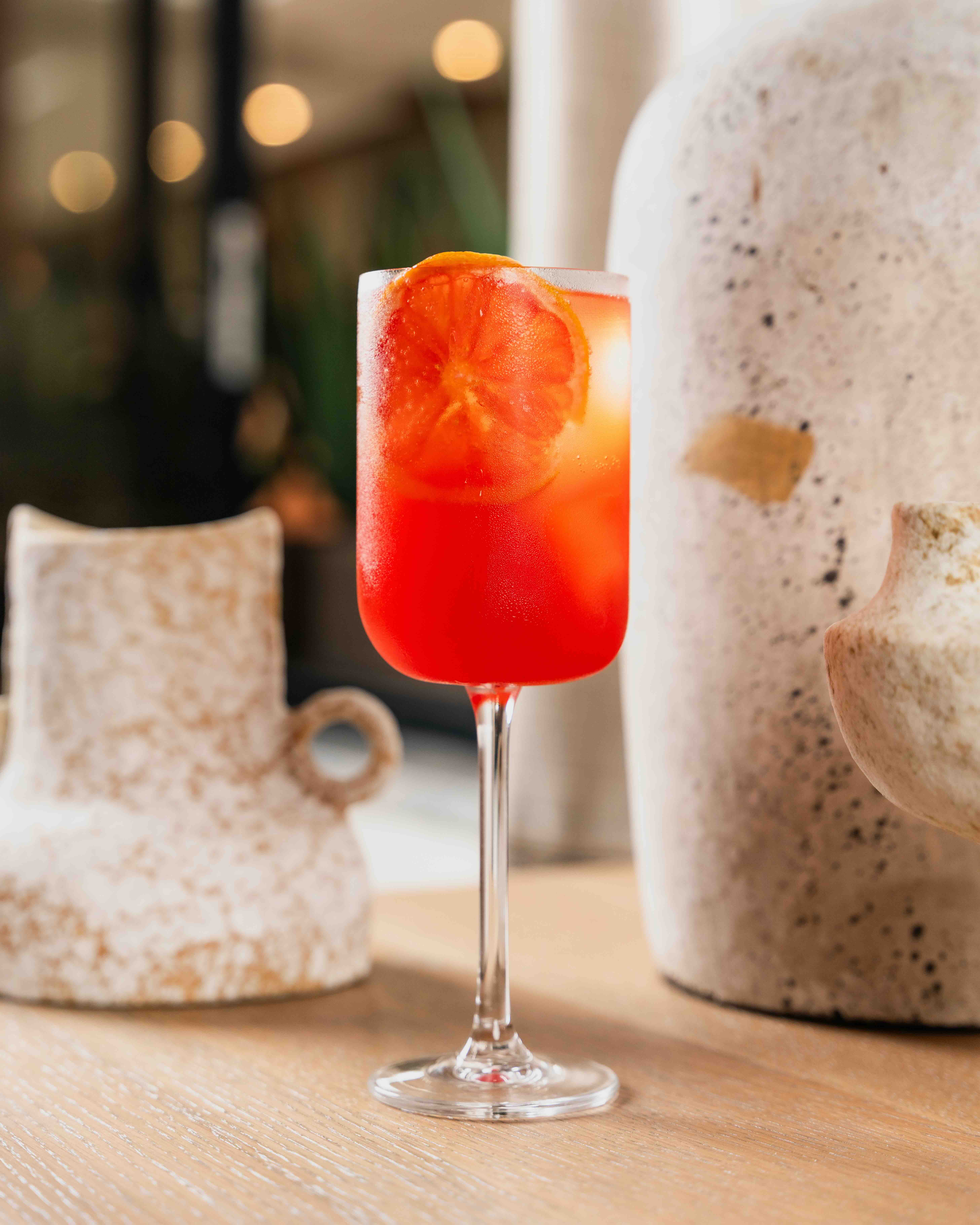 Red cocktail in a stemmed glass garnished with an orange slice on a wooden table with pottery in the background.