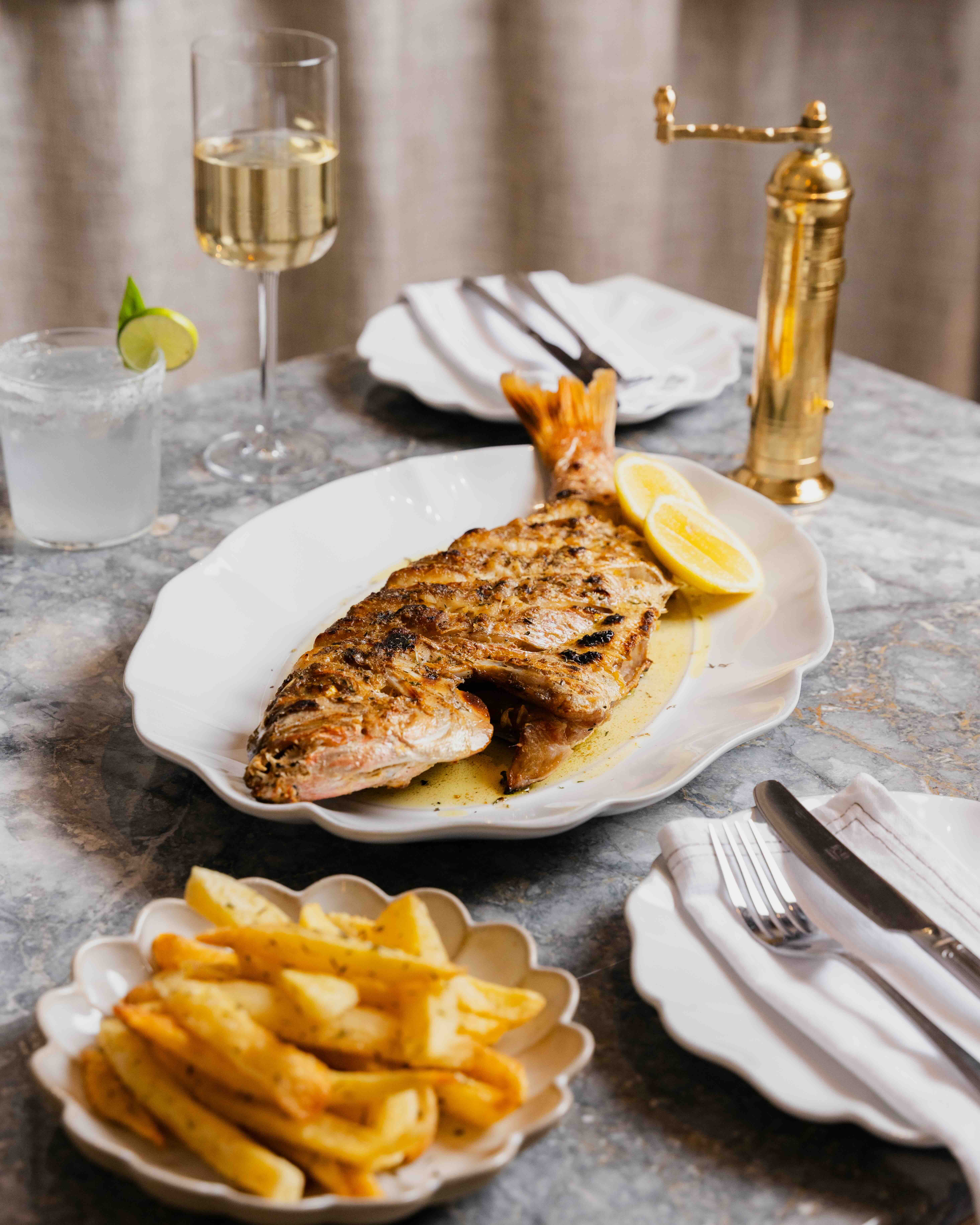Grilled whole fish with lemon wedges on a white plate served with seasoned fries and drinks on a marble table.