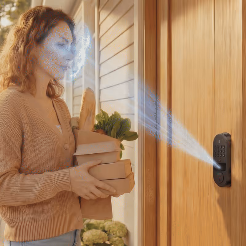Woman holding groceries using facial recognition to unlock a smart door lock on a wooden door.