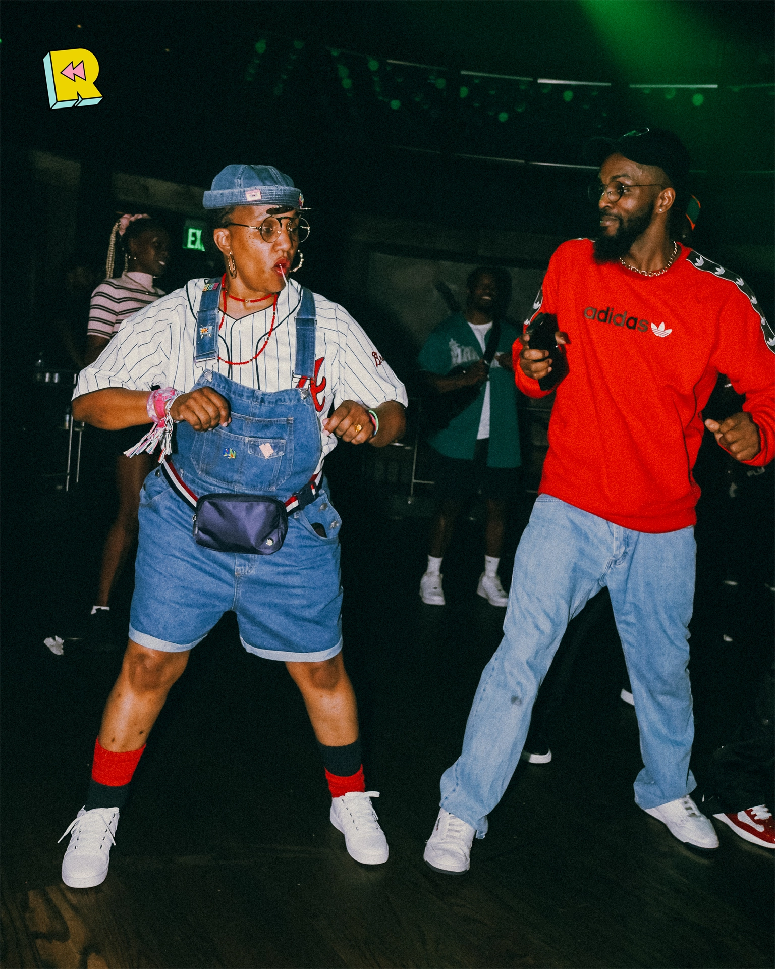 Two partygoers dancing in retro outfits at Rewind—one in denim overalls and cap, the other in a red Adidas sweatshirt.