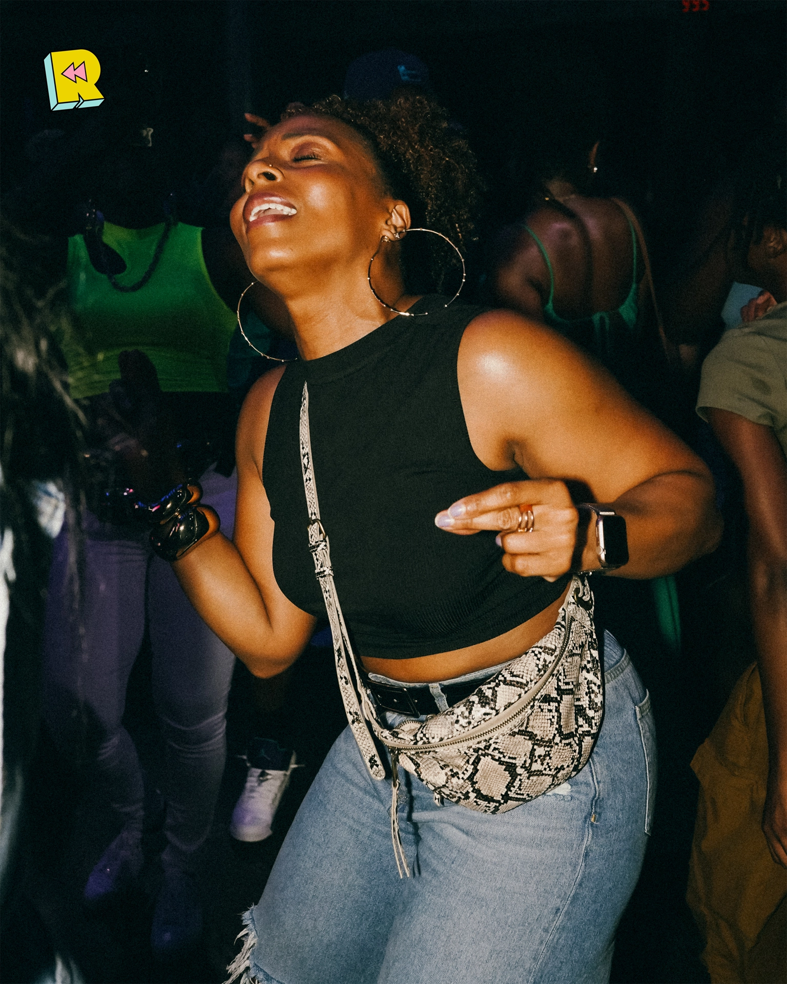 Woman dancing with joy at Rewind, wearing hoop earrings, crop top, and snakeskin fanny pack in a packed throwback party.