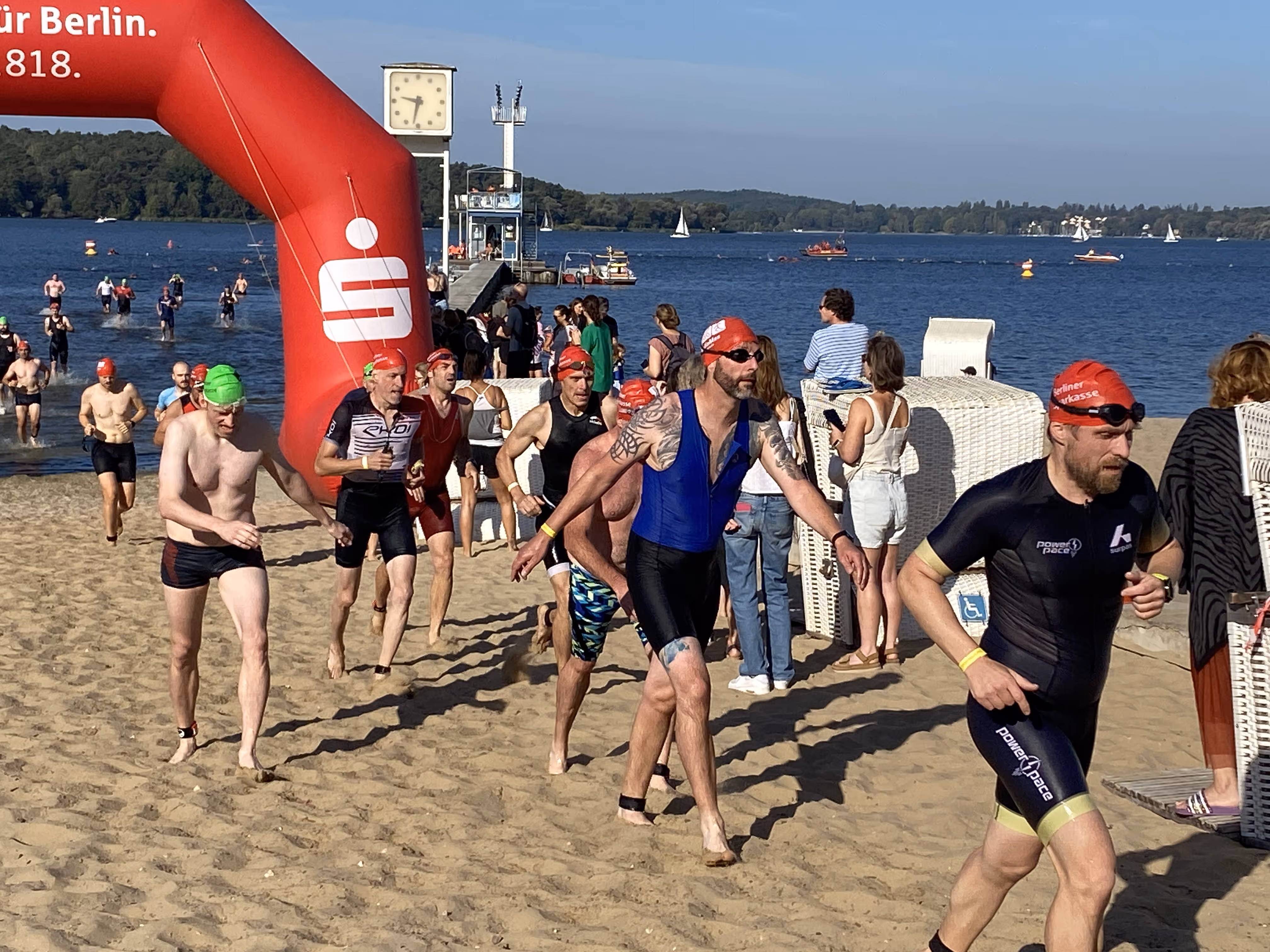 Teilnehmende des BerlinWoMan Triathlons steigen nach dem Schwimmen am Strandbad Wannsee aus dem Wasser und laufen barfuß durch den Sand Richtung Wechselzone, umgeben von Zuschauenden bei Sonnenschein.
