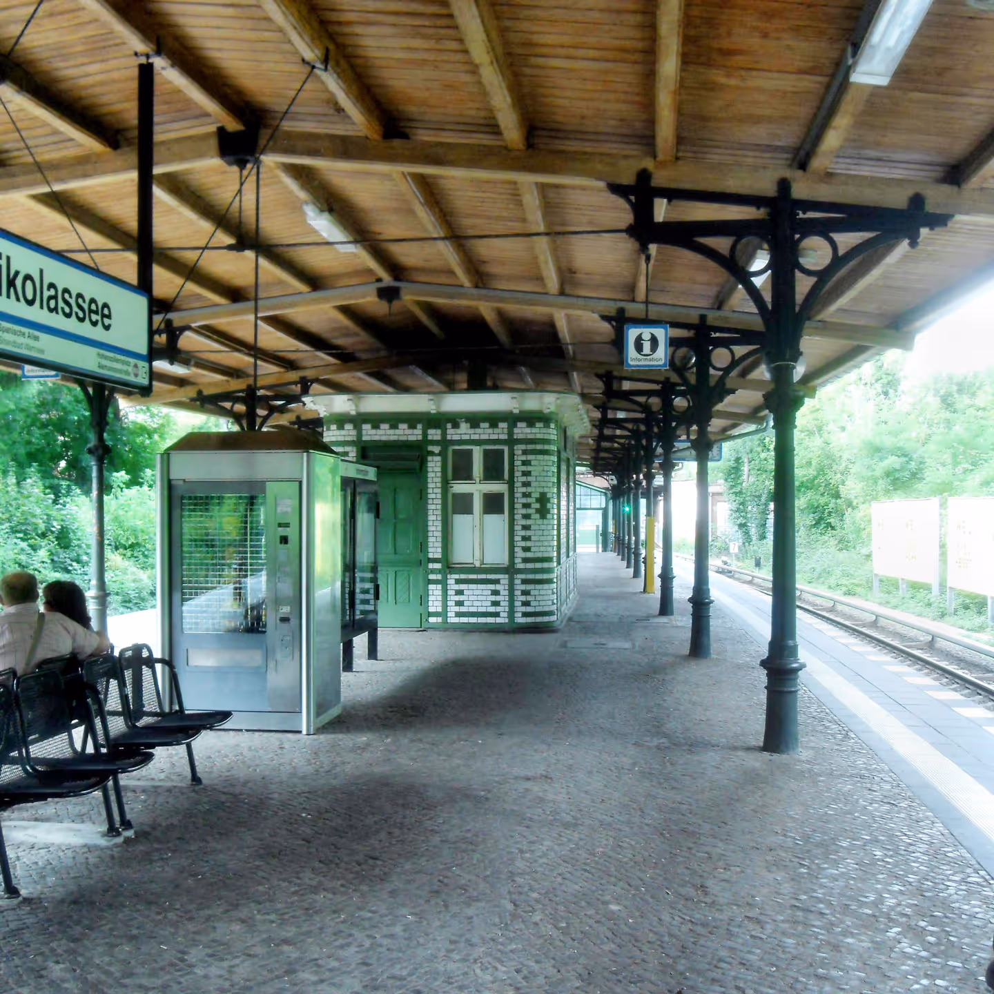 Bahnsteig des S-Bahnhofs Nikolassee mit grün-weißer Fassade und Holzdach – bequeme Anreisemöglichkeit für Zuschauer*innen des BerlinWoMan Triathlons.