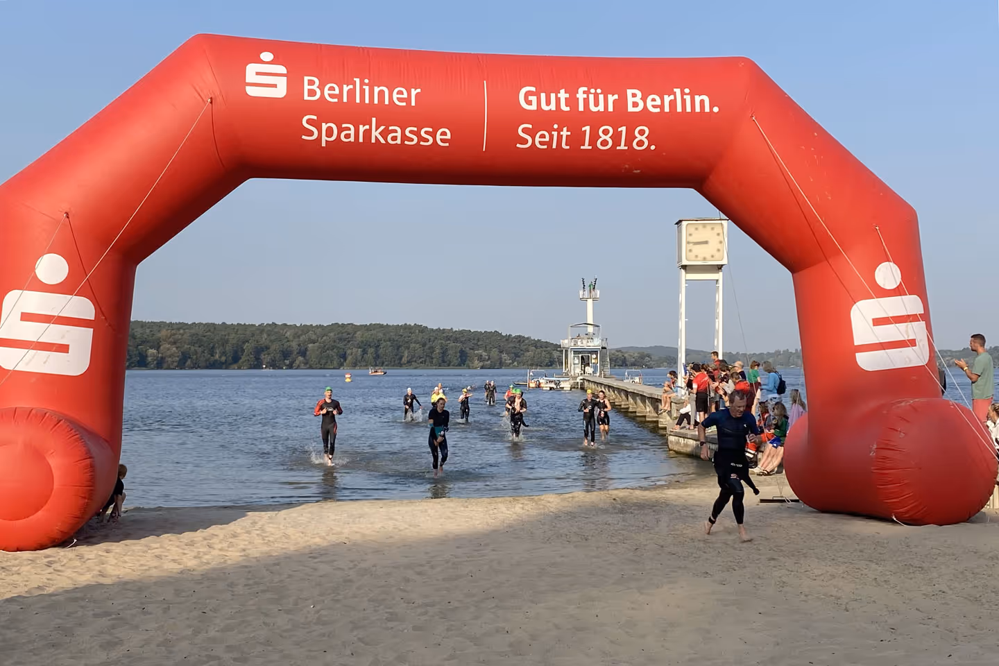 Teilnehmer in Neoprenanzügen laufen aus dem Wasser an einem Strand unter einem roten Berliner Sparkasse Torbogen.
