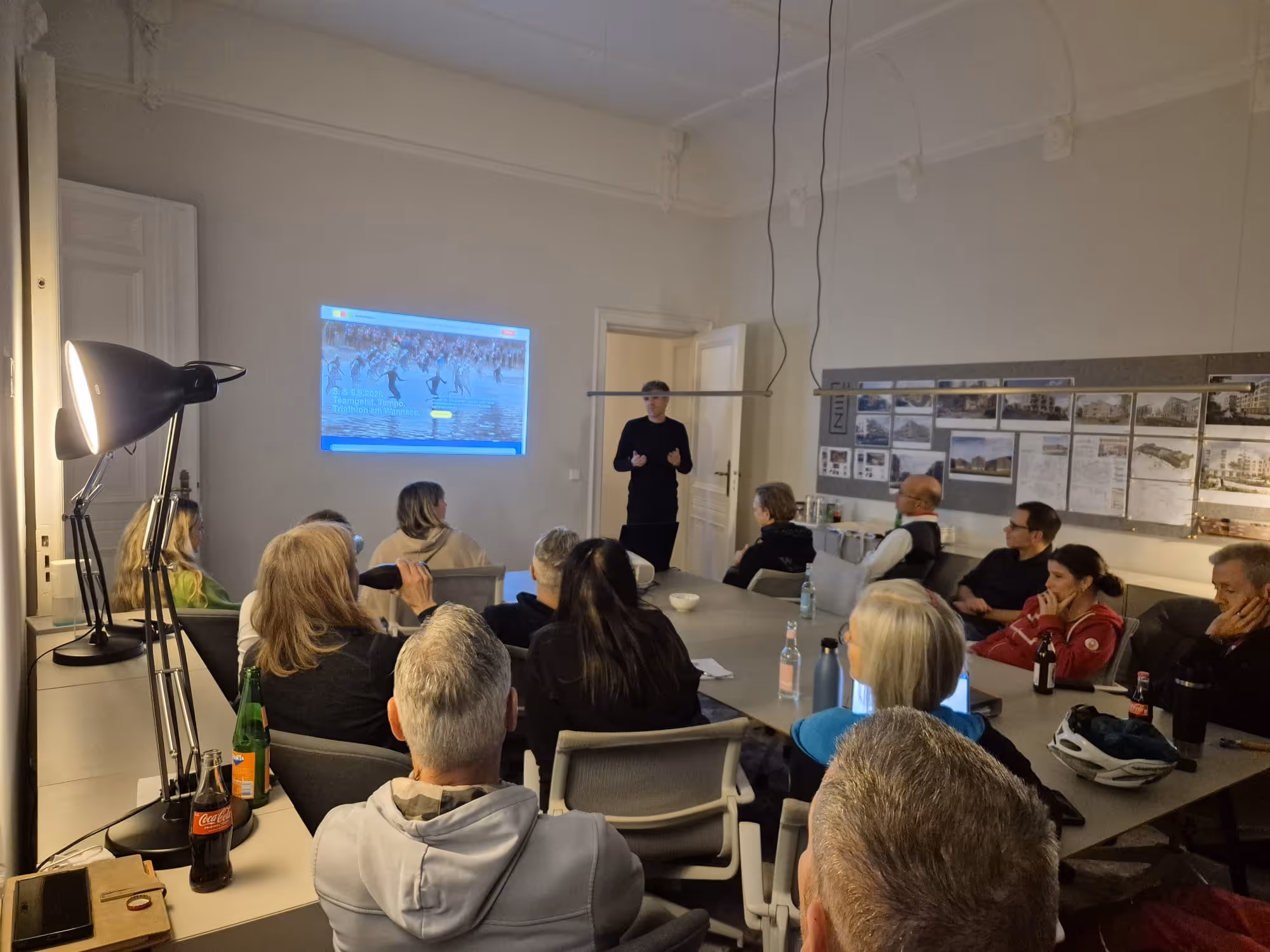 Die Tophelfer des BerlinWoMan 2026 sitzen an einem Konferenztisch und hören Silvio zu, der vor einer Wand mit einem projizierten neuen Website steht.