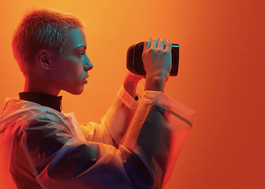 Futuristisch wirkender junger Mann mit Cyberbrille und Kamera, in orangem und violettem Licht inszeniert – technikaffine Bildsprache im Sci-Fi-Stil.
