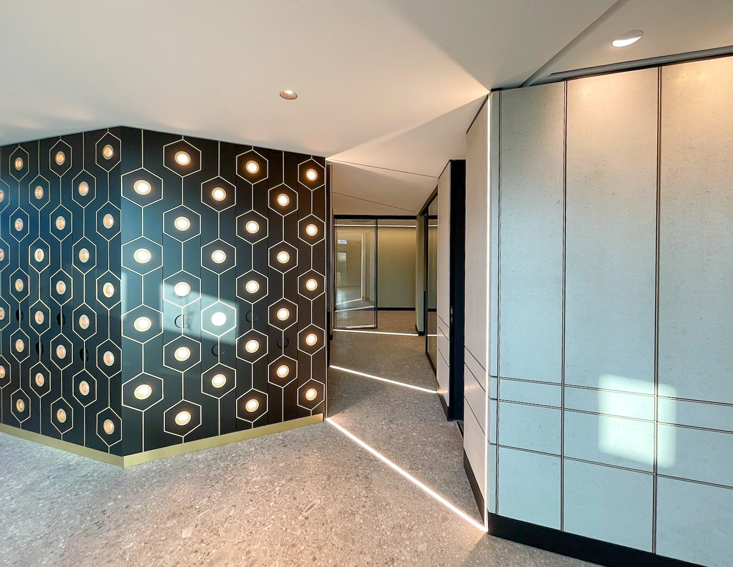 A modern hallway with patterned black wall panels illuminated by geometric lights and soft floor lighting.