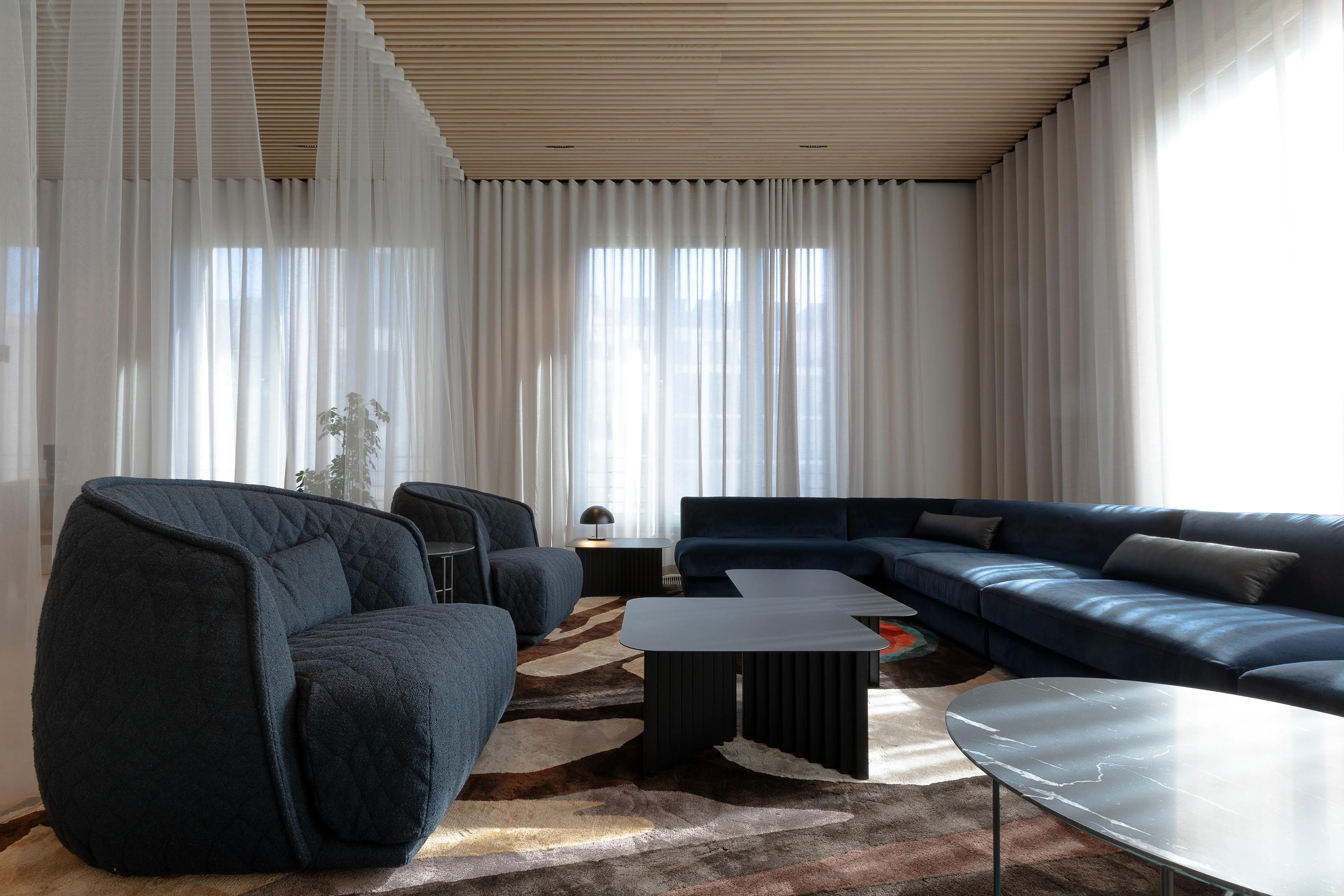 A modern living room with dark blue sofas, sheer white curtains, a wooden ceiling, and a mix of minimalist coffee tables.
