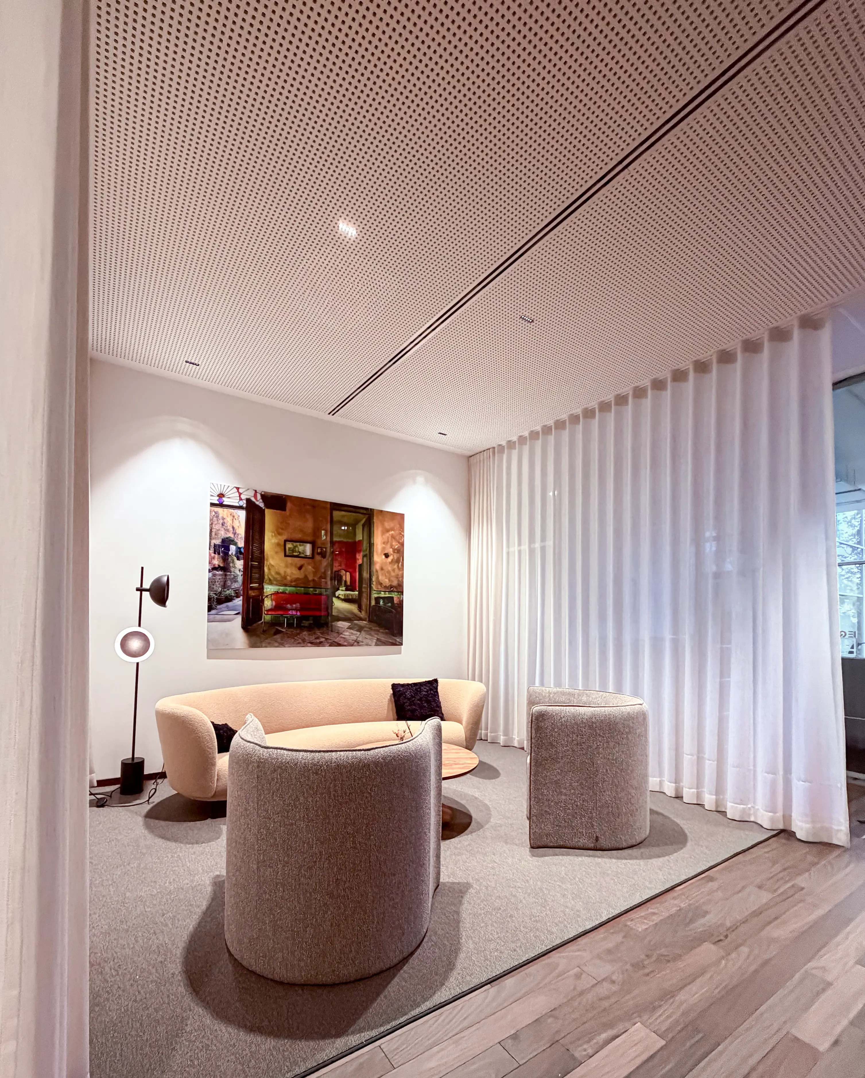 A cozy contemporary lounge with beige furniture, soft lighting, a colorful wall painting, and floor-to-ceiling white curtains.