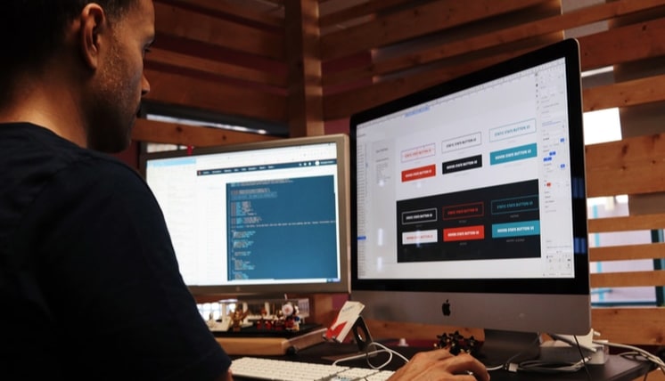 Person working at a desk with dual monitors, one displaying code and the other showing a UI design interface, in a modern wood-paneled office environment.