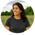 Profile photo of a woman with long dark hair wearing a dark t-shirt, smiling while standing outdoors in a grassy field with trees in the background.