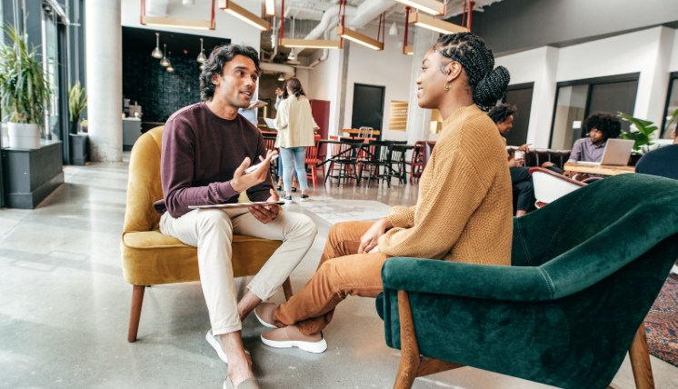 wo people seated in a modern office setting engaged in a conversation, likely a recruiter screening interview. One holds a tablet while listening attentively to the other.