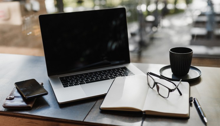 Laptop, notebook with glasses, smartphone, and a coffee cup arranged on an outdoor table, suggesting a focused work or study environment for a TPM role introduction.