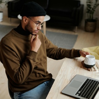 Man wearing glasses and a beanie, sitting at a table with a tablet and holding a coffee cup, appearing focused and thoughtful.