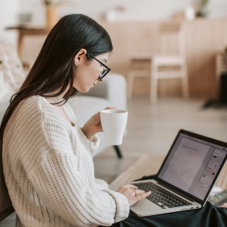 Woman in glasses and a cozy sweater working on a laptop while holding a coffee mug, seated in a warm, minimal indoor setting.