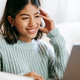 Smiling woman wearing a light knit sweater, looking at a laptop screen with her hand resting on the side of her head, suggesting engagement in an online activity.