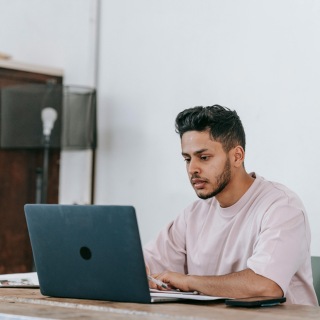 Focused man working on a laptop in a minimal indoor setting, wearing a light pink sweatshirt and seated at a wooden desk.