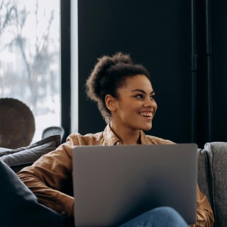 Smiling woman sitting indoors with a laptop on her lap, looking to the side, appearing engaged and relaxed in a cozy setting