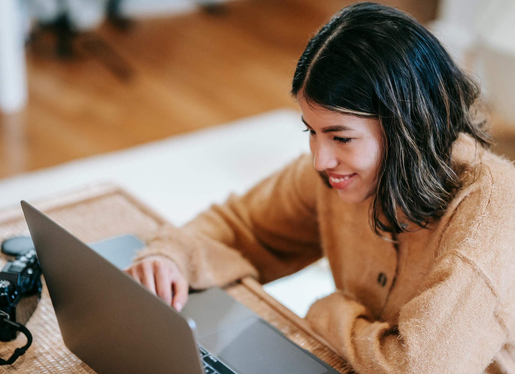 Smiling woman in a brown sweater working on a laptop at home, seated at a table with a woven placemat and a camera nearby.