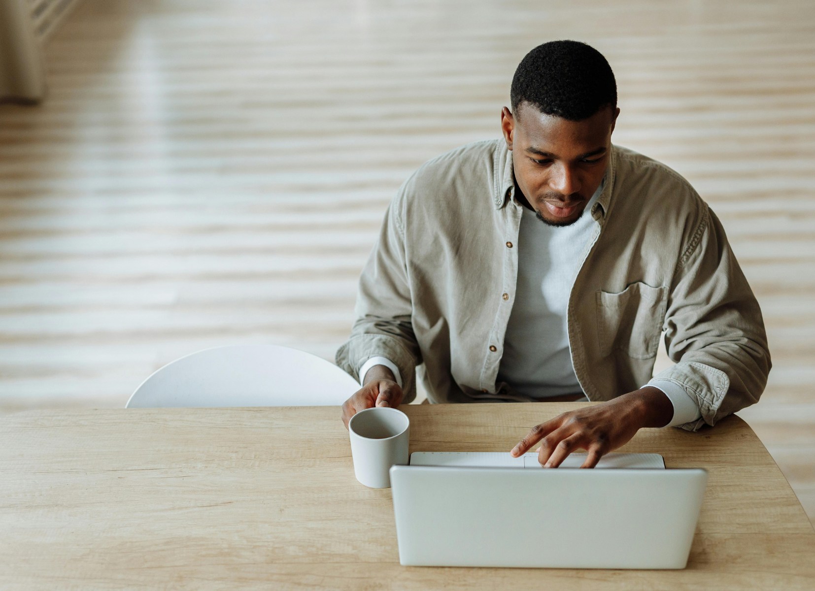Man sitting at a wooden table using a laptop while holding a white coffee mug, focused on his work in a bright, minimalistic space.