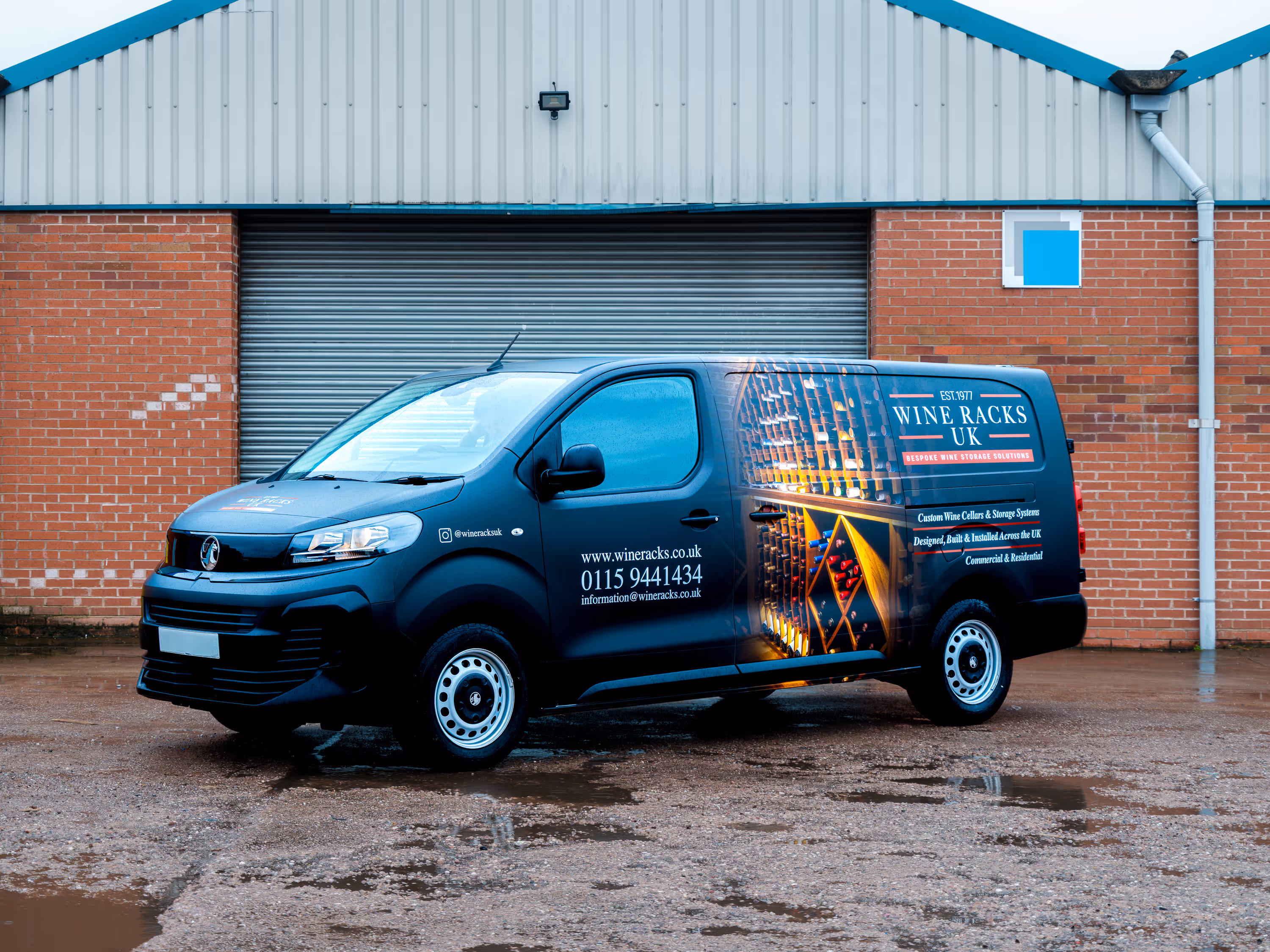 Black delivery van with 'Wine Racks UK' branding parked in front of a brick building with a closed metal shutter.