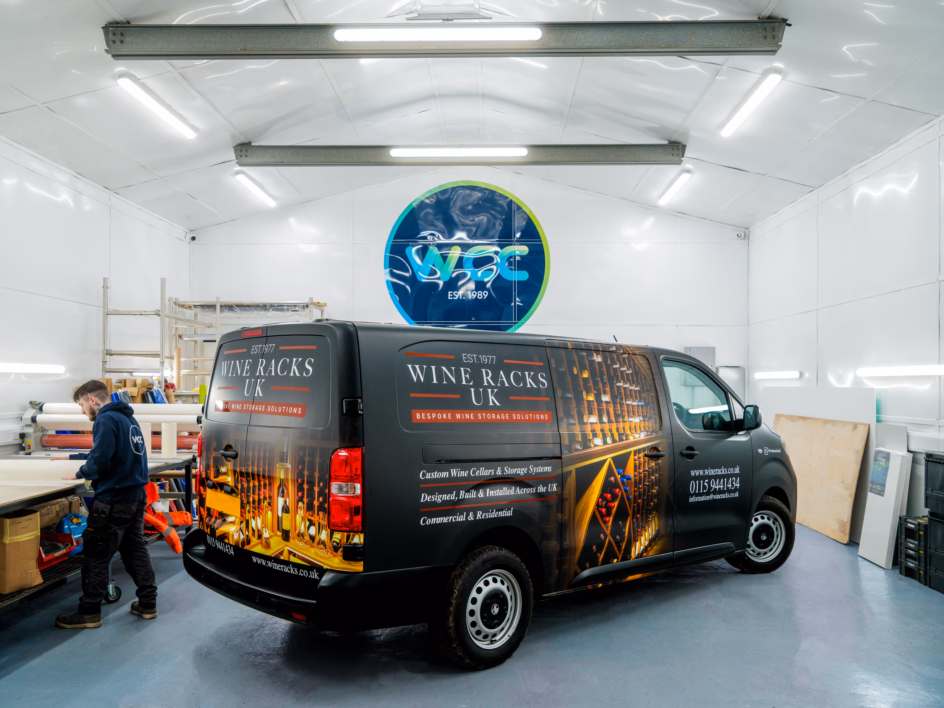Black van inside a workshop with Wine Racks UK branding and wine storage images, a man working nearby.