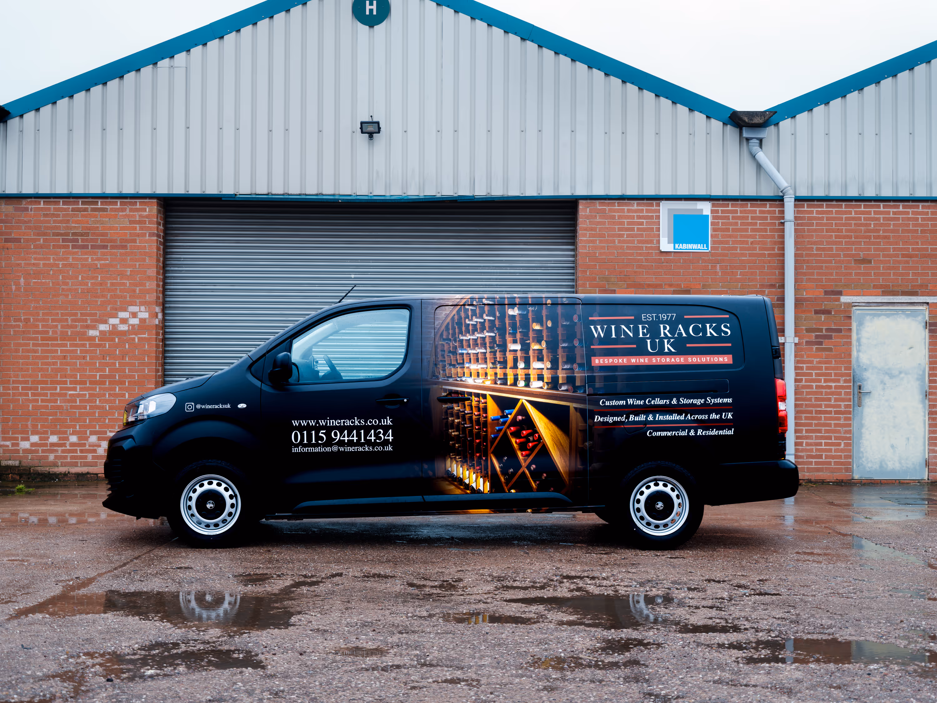 Black van with wine racks UK advertisement featuring wine bottles, contact details, and services parked in front of a brick building with a closed shutter.