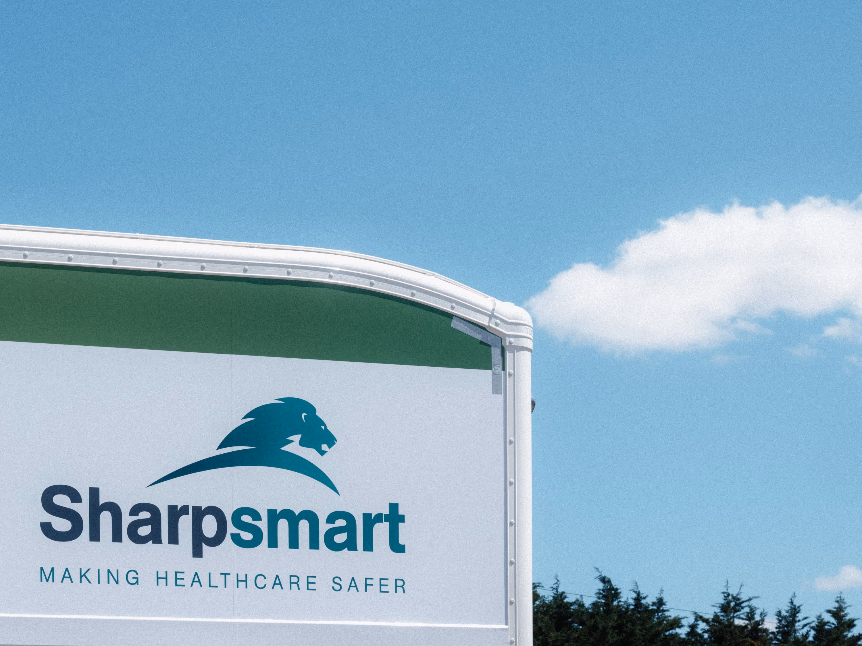 Side of a SharpSmart trailer with a lion logo and the tagline Making Healthcare Safer against a blue sky with a cloud and trees below.