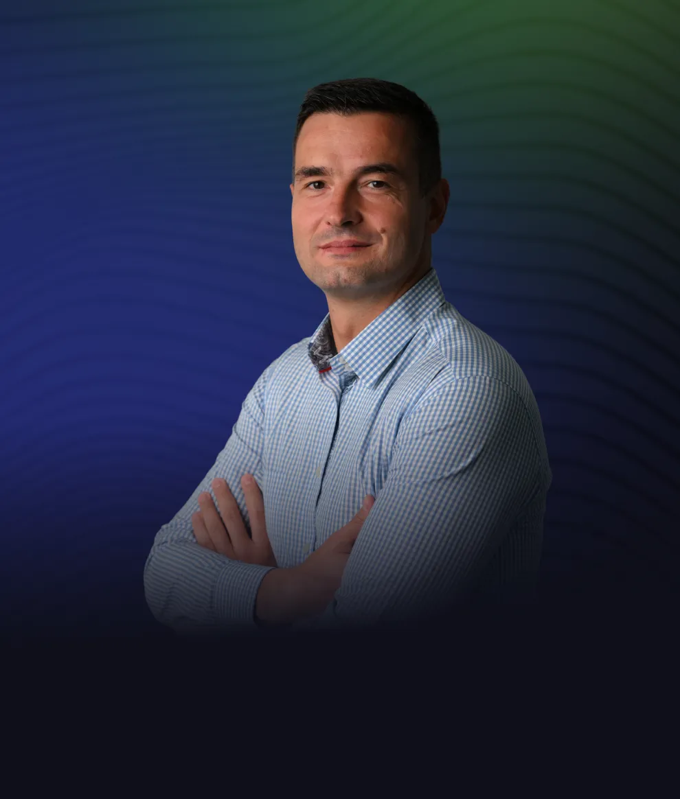 Man with short dark hair wearing a light blue checkered shirt, standing with arms crossed and smiling slightly against a blue and green gradient background.