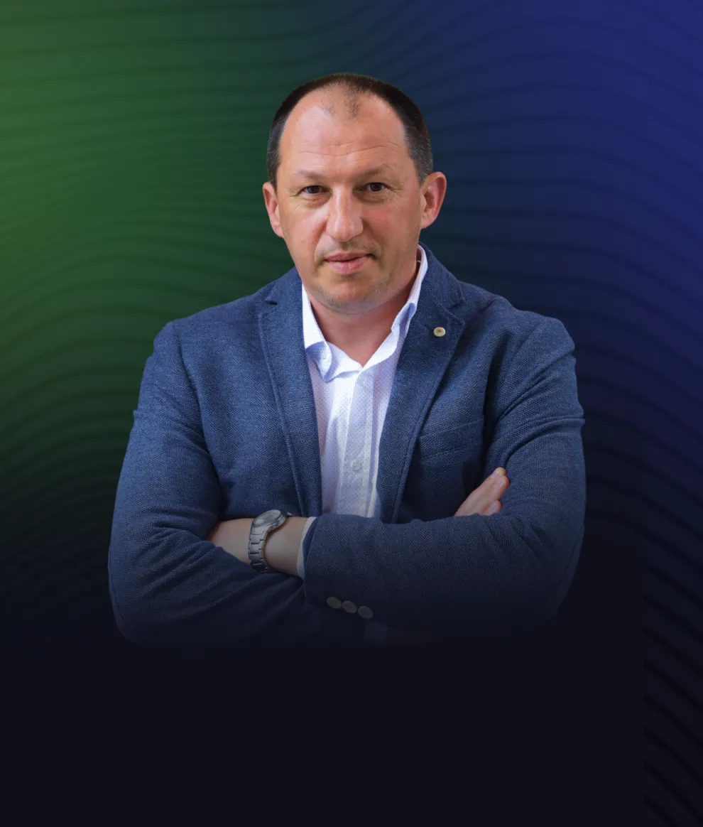 Man with short dark hair wearing a blue blazer and white shirt with arms crossed, against a dark blue and green gradient background.