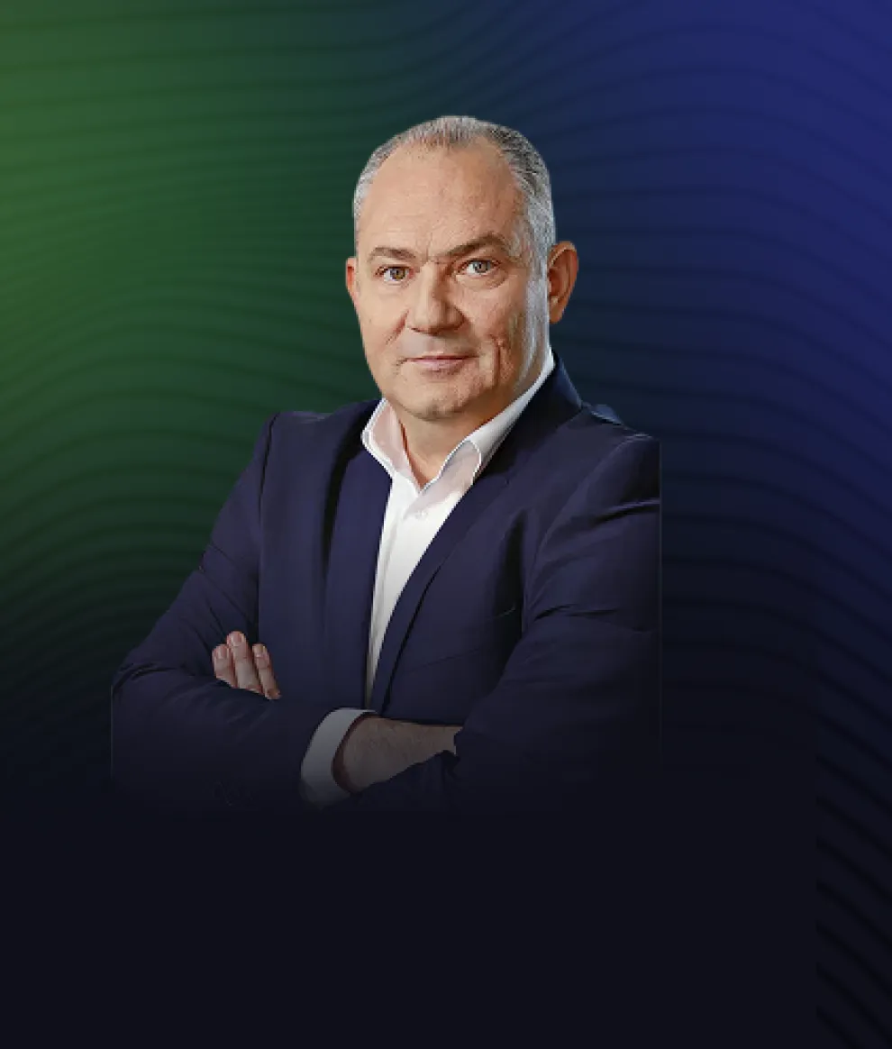 Portrait of a middle-aged man with gray hair, wearing a dark suit and white shirt, arms crossed, against a blue and green gradient background.