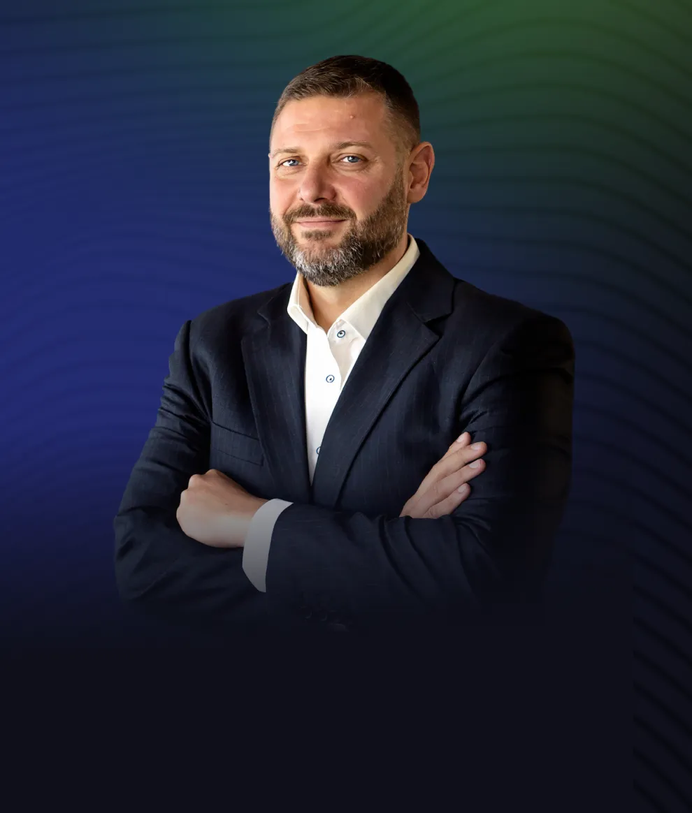 Man with beard and short hair wearing a dark suit and white shirt, standing with arms crossed against a blue and green gradient background.