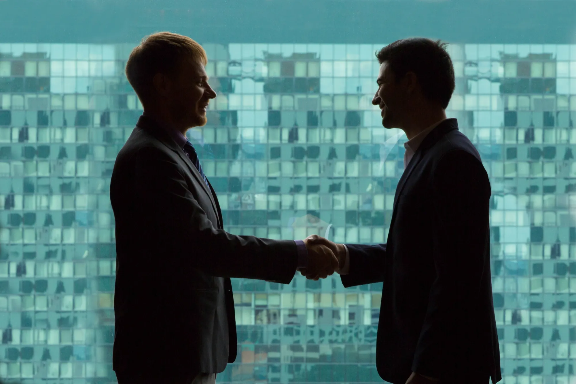 Two men in suits shaking hands and smiling in front of a large window with a grid-patterned cityscape view.