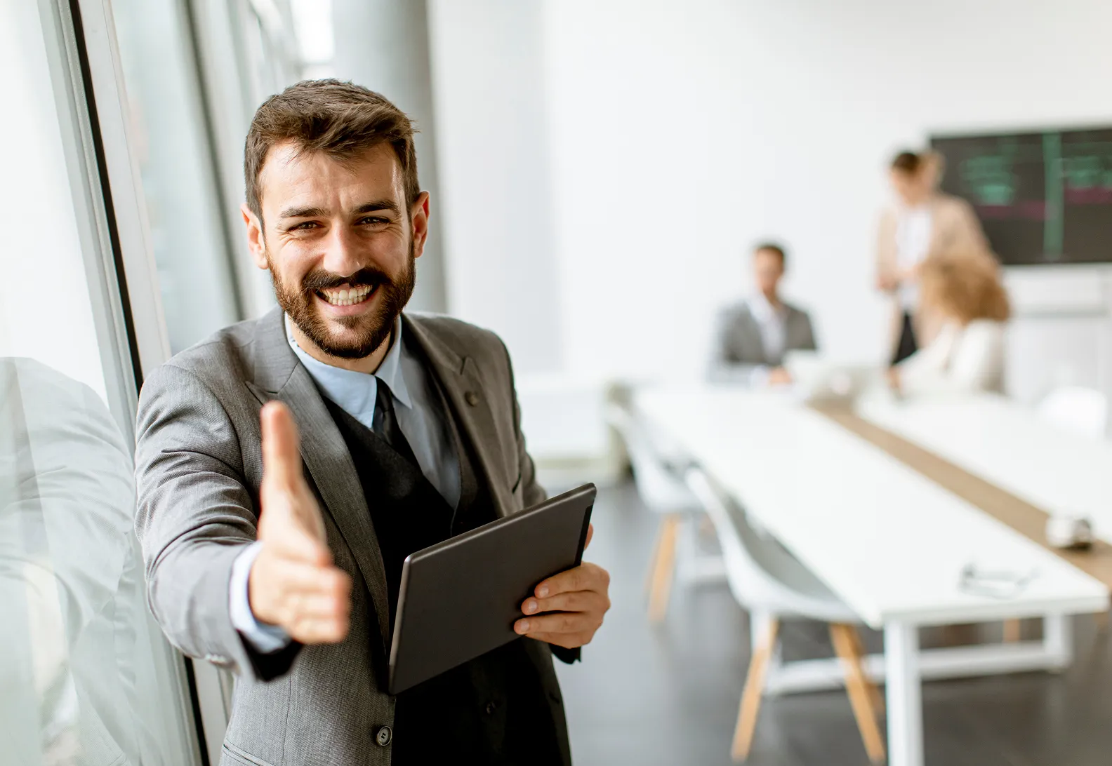 Smiling businessman in a gray suit offering a handshake while holding a tablet with colleagues working in the background.