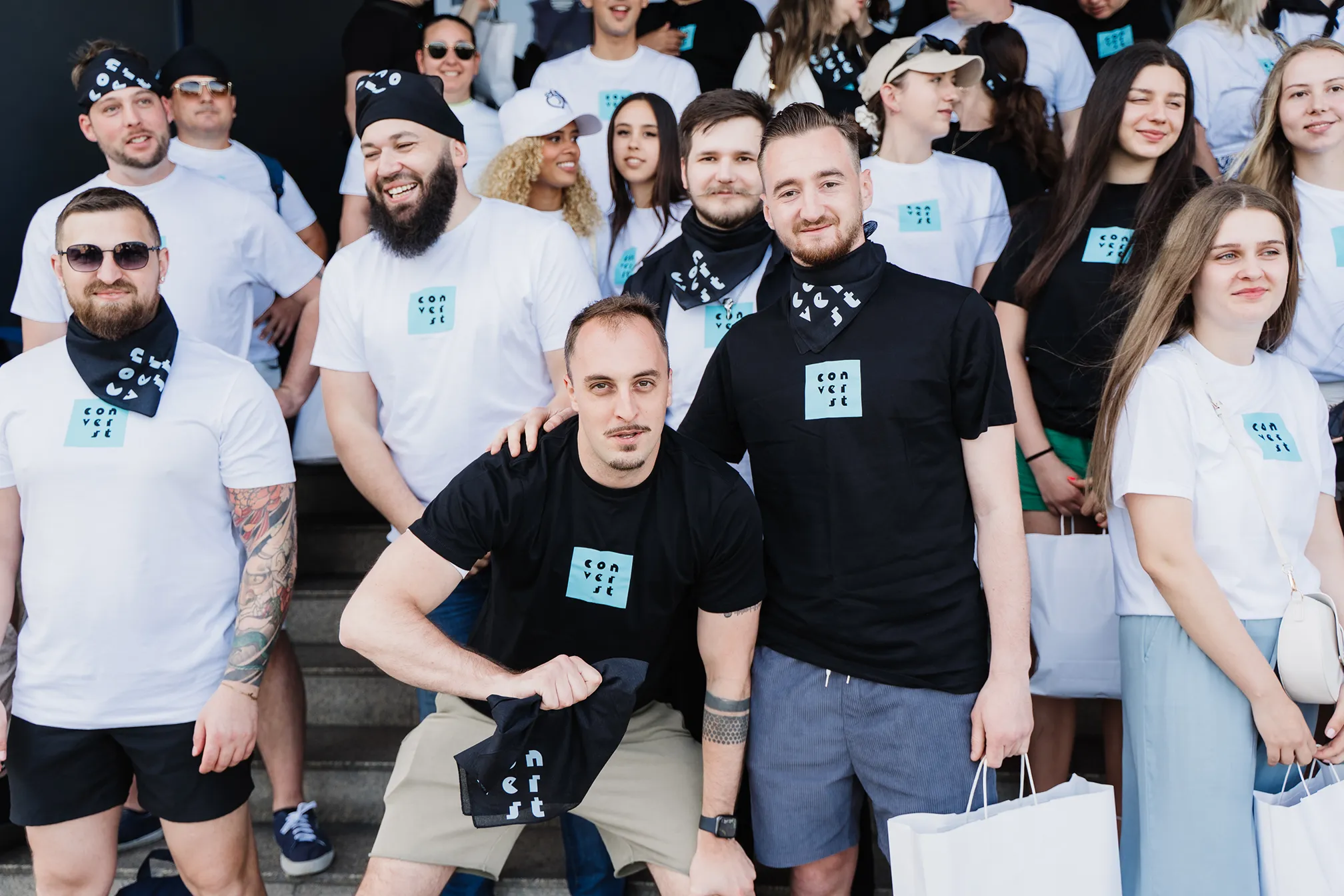 Group of smiling young adults wearing white and black t-shirts with 'converst' logo, posing together on outdoor steps during daytime.