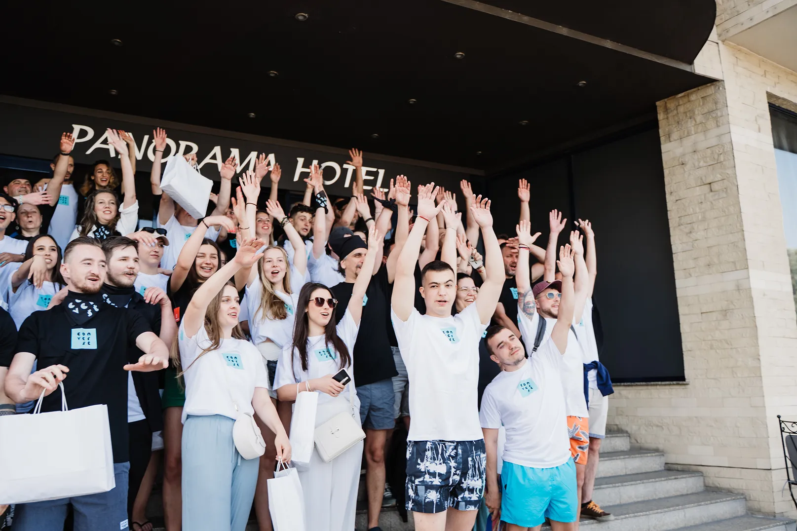 Group of young people in matching white and black Converse t-shirts raising their hands while standing on hotel steps outside Panorama Hotel.