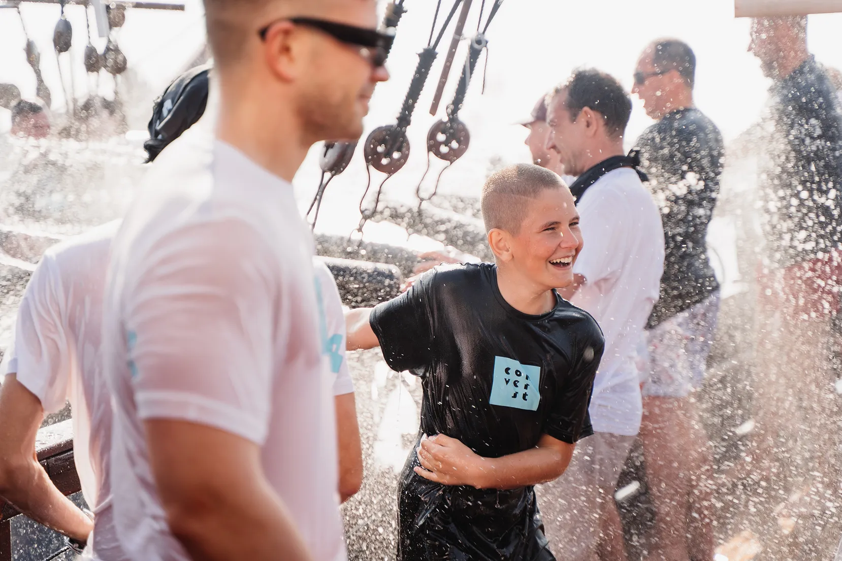 Smiling boy in a black wet t-shirt laughing while being splashed with water among a group of people wearing white and black shirts on a boat.
