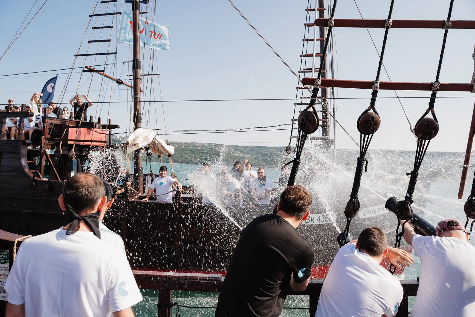 People on two pirate-style ships splashing water at each other on a sunny day near a coastline.