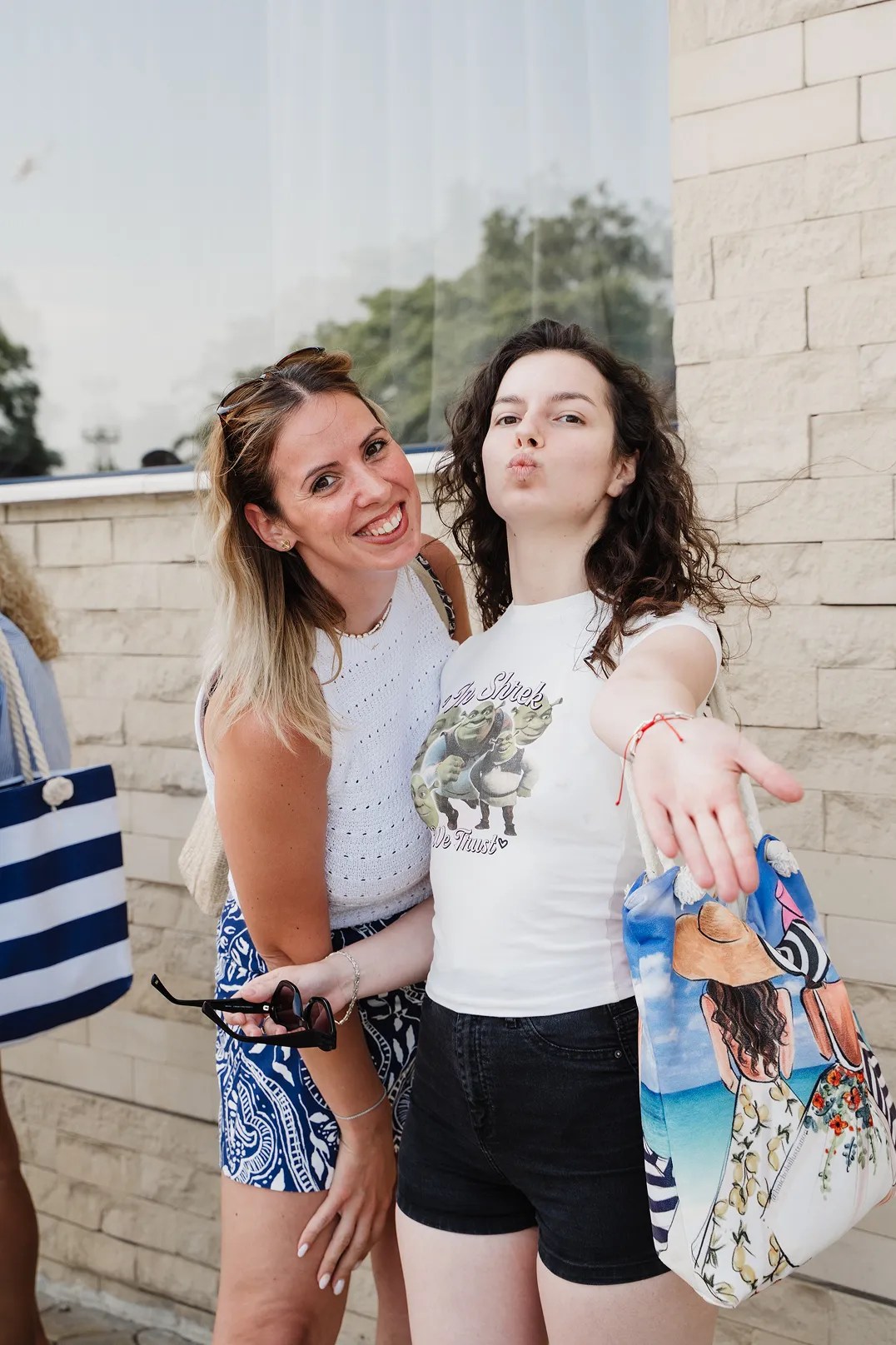 Two young women posing outdoors, one smiling at the camera and the other making a kissing face while holding a colorful beach bag.