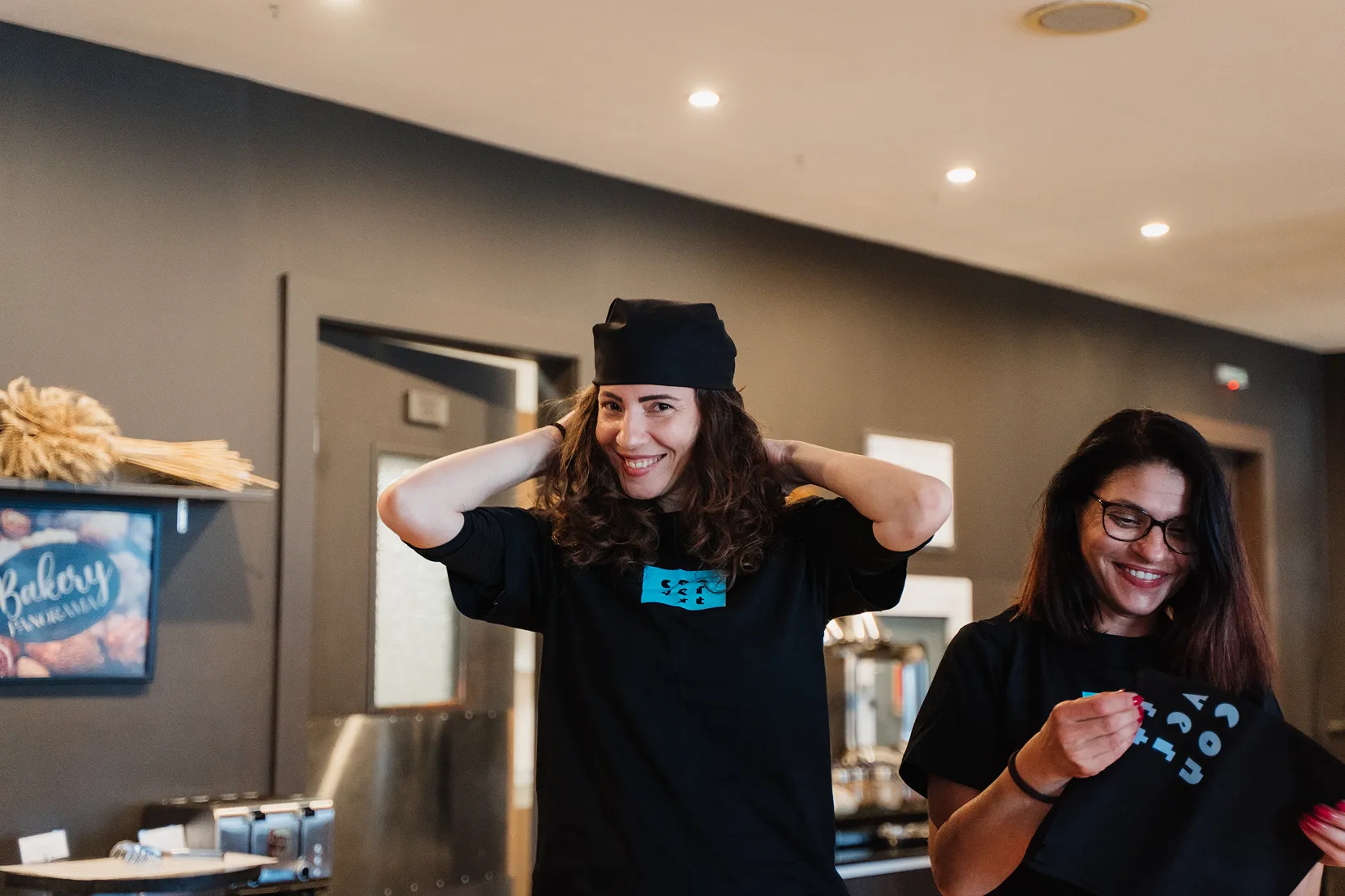 Two smiling women in black shirts and headwear inside a bakery or café setting with bakery decor in the background.