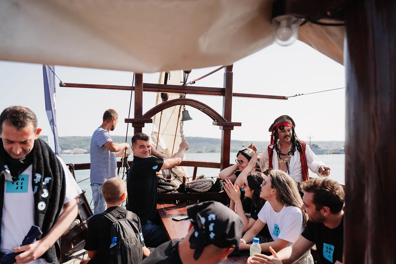 Group of people enjoying a wooden boat ride with one man ringing a bell and another dressed as a pirate.