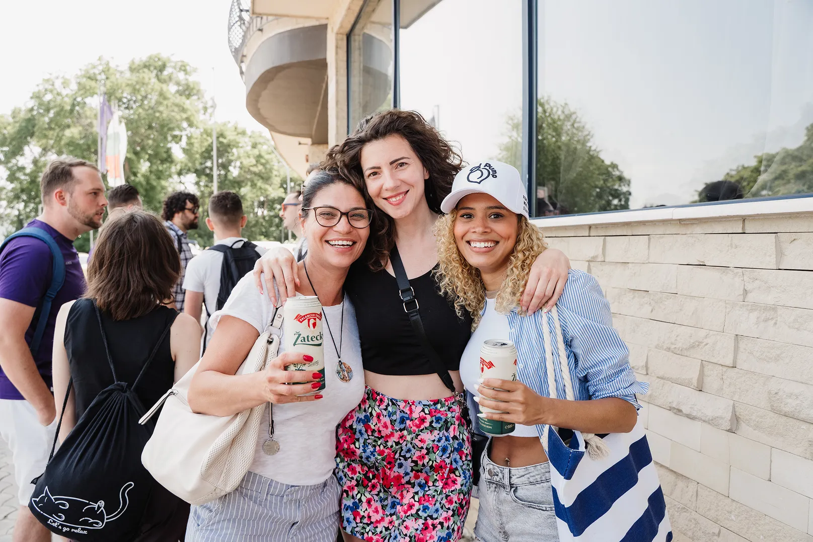 Three smiling women with arms around each other, holding drinks outdoors near a building.